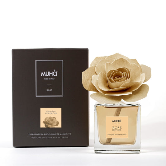 Profumatore d'ambiente Muhà Rose Diffuser Vaniglia e Ambra Pura in flacone squadrato di vetro trasparente con essenza dorata, etichetta centrale quadrata e diffusore artigianale a forma di rosa in legno ocra chiaro, Tutto Chic.