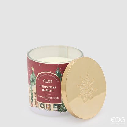 Candela natalizia profumata EDG in grande bicchiere rosso satinato decorato "Christmas Hamlet", con cera bianca e coperchio lucido in metallo dorato con fiocco di neve a rilievo.