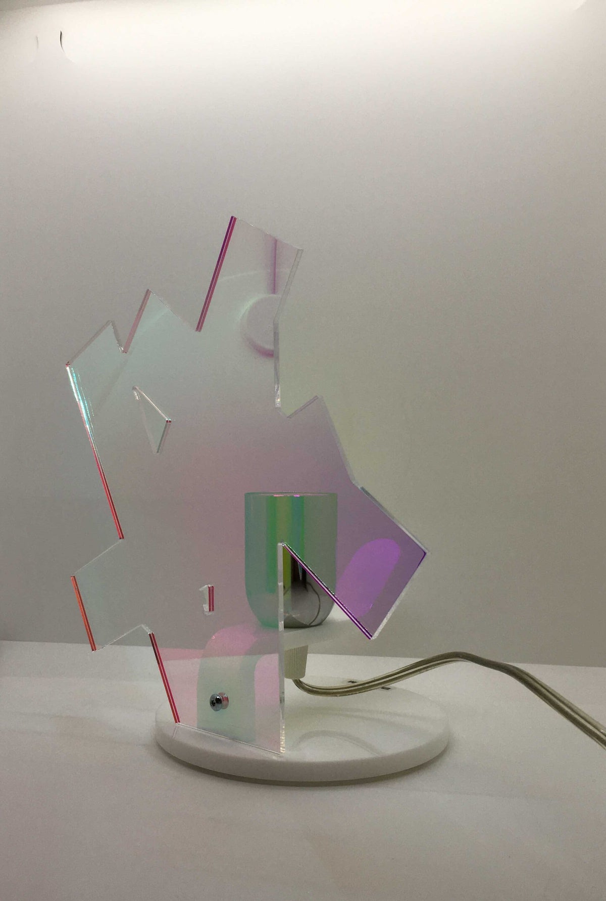 Lampada da tavolo in metacrilato dalla colorazione particolare flash (effetto specchio iridescente). Base e supporto in metacrilato bianco e portalampadina in metallo cromato. Si può applicare qualsiasi lampadina con attacco filettato E 27 (non inclusa). Dimensioni: cm 23 x 23 x 15 p. In negozio e online su tuttochic.live