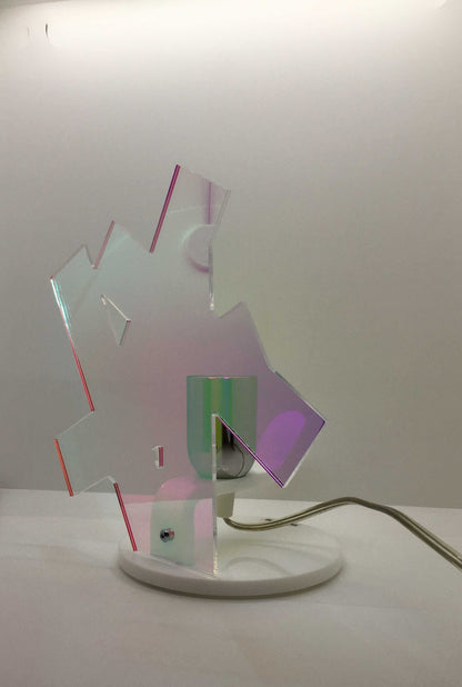 Lampada da tavolo in metacrilato dalla colorazione particolare flash (effetto specchio iridescente). Base e supporto in metacrilato bianco e portalampadina in metallo cromato. Si può applicare qualsiasi lampadina con attacco filettato E 27 (non inclusa). Dimensioni: cm 23 x 23 x 15 p. In negozio e online su tuttochic.live