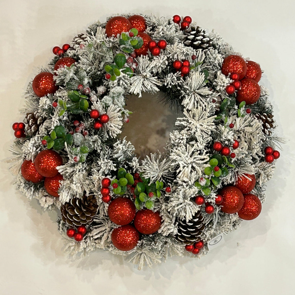 Realizzata con 135 rametti di pino artificiale innevato in pvc. Decorata con rametti artificiali di Bosso con bacche, pigne decorate, bacche in vetro di colore rosso e palline in plastica rosso glitter Diametro cm 60 circa. In negozio e online su tuttochic.live