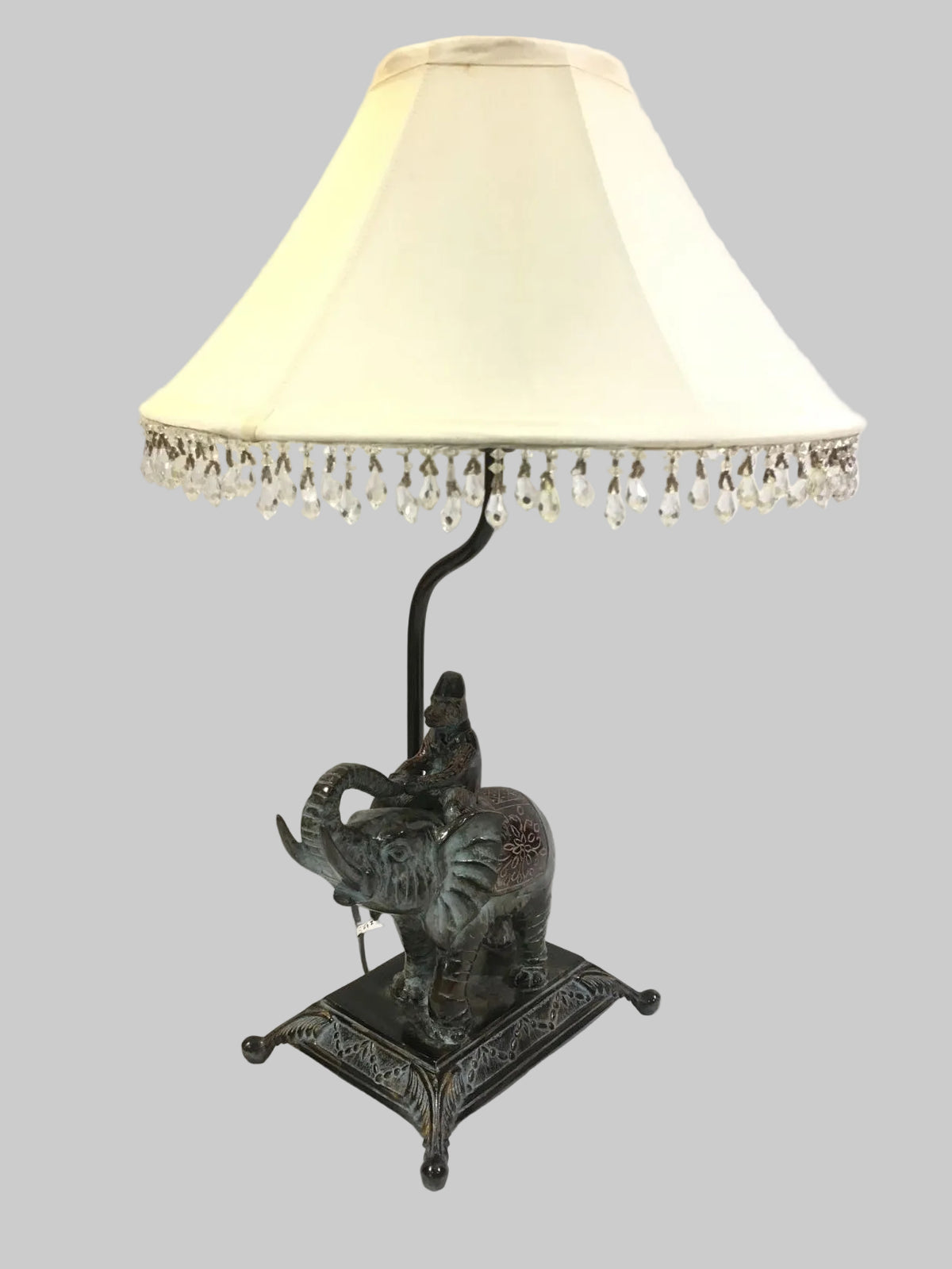 Lampada d'arredo in metallo effetto bronzo di colore verde con paralume in tessuto di colore beige chiaro decorato con strass in acrilico trasparente. Alimentazione: lampadina attacco E27 max 60 W Dimensioni: base cm. 21 x 16 x 55 H - paralume ø 35. In negozio e online su tuttochic.live
